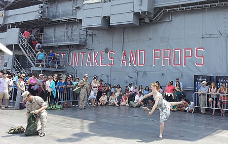 Woman in flower-print dress dancing on a battleship cruiser with a crowd watching her and a man in miltary uniform unpacking items from a green bag.