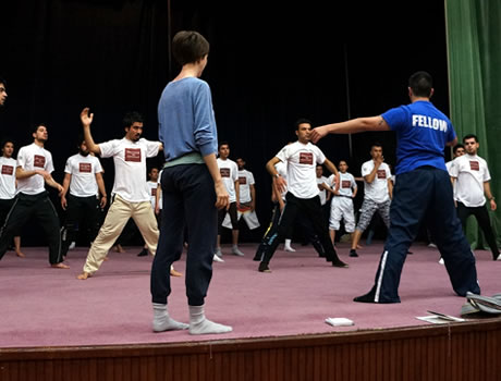 A man in blue shirt with 'Fellow' on the back, with a woman in a blue shirt next to him, directing dancers in white shirts.