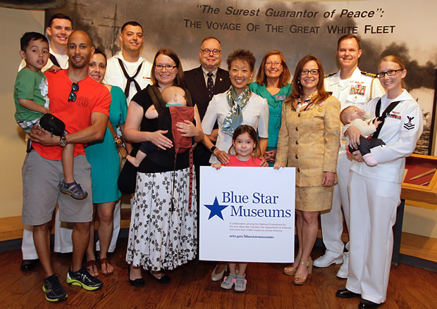 Large group of men, women, and children posing for a group photo, Child in the front middle holding the Blue Star Museums sign
