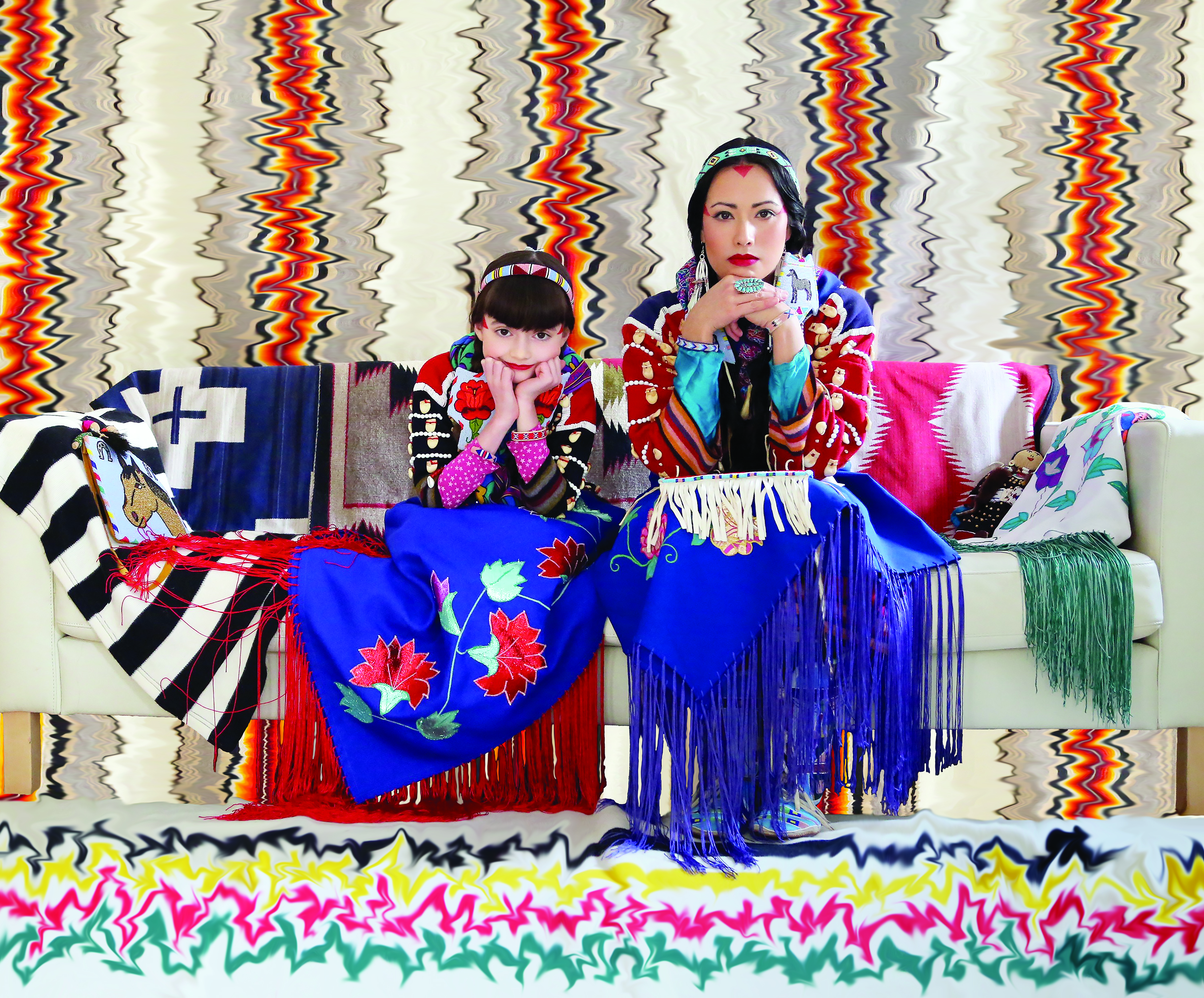 Woman and young girl dressed in Native garb sitting on a couch before a background of dizzying patterns.