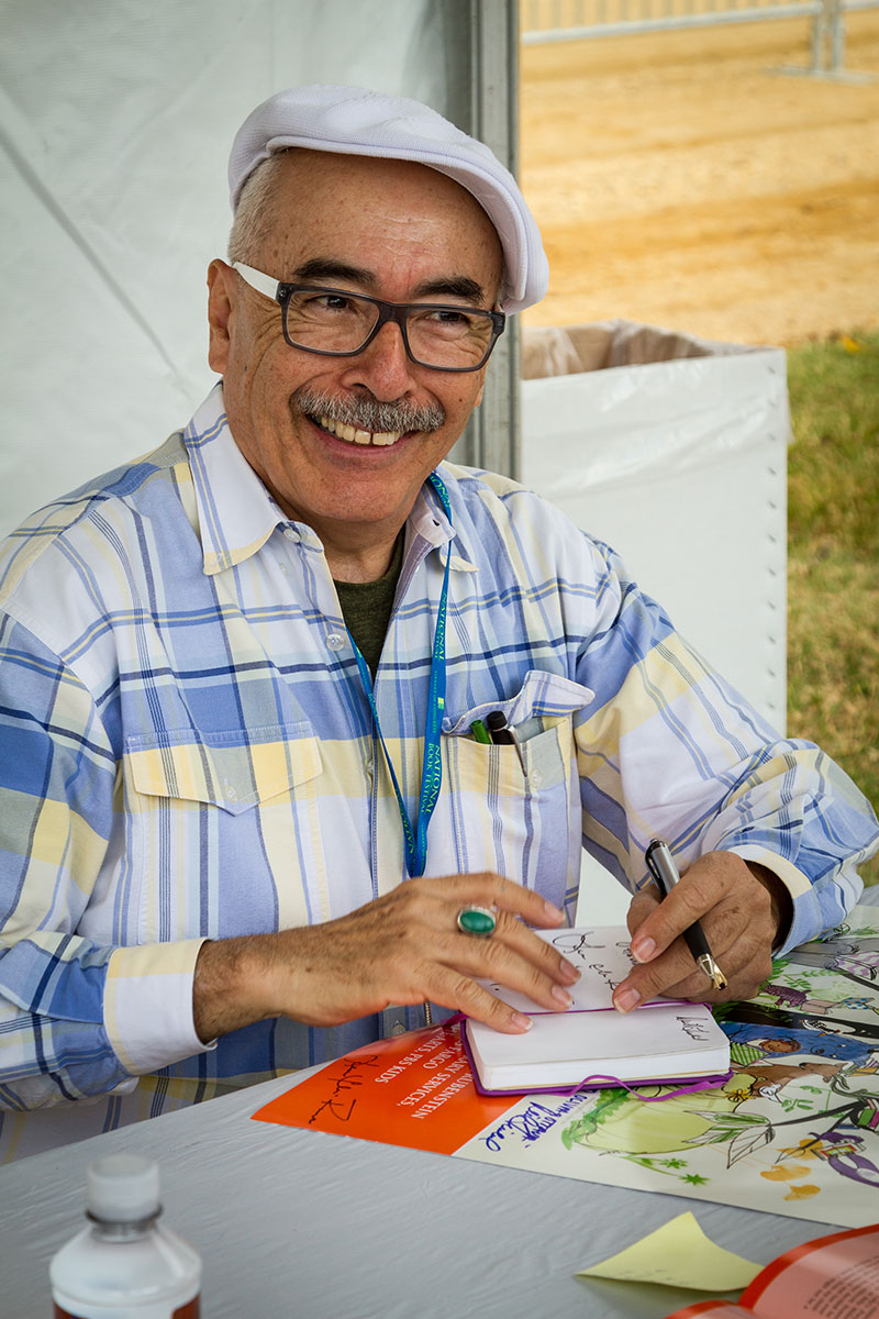 Juan Felipe Herrera sitting down at a table with a pen in his hand and looking up smiling.