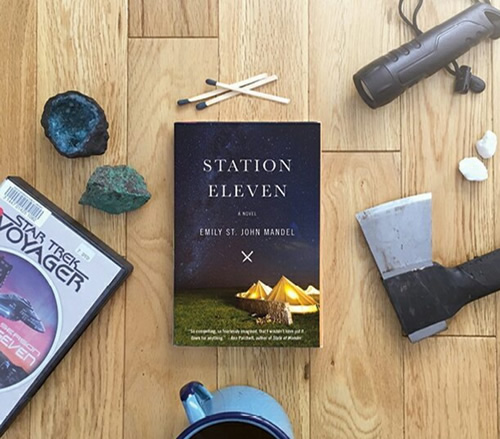  A copy of Station Eleven, surrounded by various survival tools, a coffee mug, and a DVD copy of “Star Trek Voyager.”
