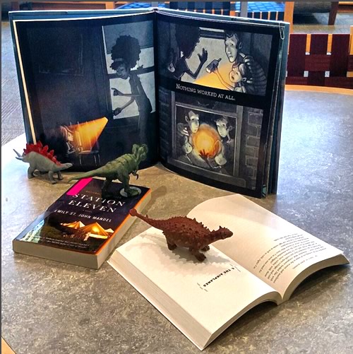 Display of Station Eleven and companion books for young readers with dinosaur figurines.