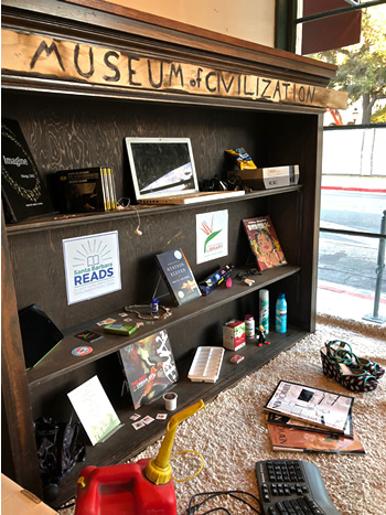 Shelf labeled “Museum of Civilization” containing various items like an old laptop, an old Nintendo, copies of the New Yorker, and an ice cube tray.