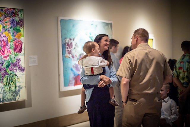 a  man and a woman holding a child talk to each other against a background of colorful paintings