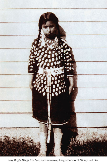 Black and white photo of a young Native girl standing in front of a wooden house. 