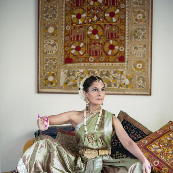 A woman in classical Indian dress seated amongst colorful pillows strikes the pose of the goddess Devi