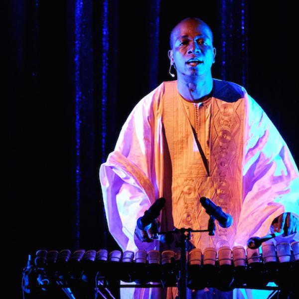 A man stands on a dark stage playing the balafon and singing.