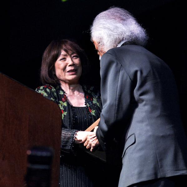 Japanese woman shaking hands with white-haired man in suit on stage. 