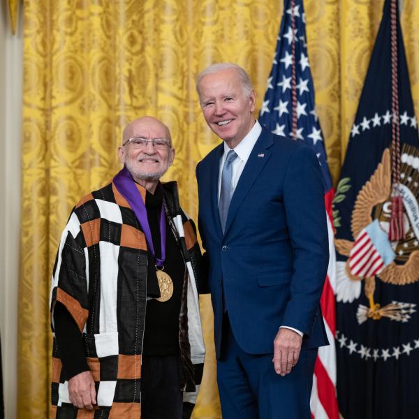 Older white male in blue suit posing with older Hispanic man wearing a black-brown-white patchwork jacket in front of flags and gold curtain.