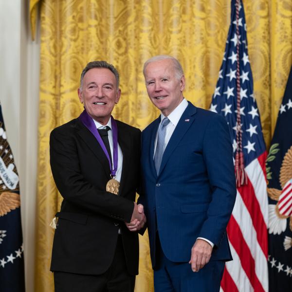 Older white male in blue suit posing with man in black suit in front of flags and gold curtain.