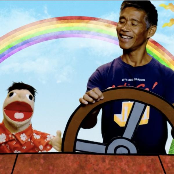 A smiling man sits next to a puppet while they drive in an animated car