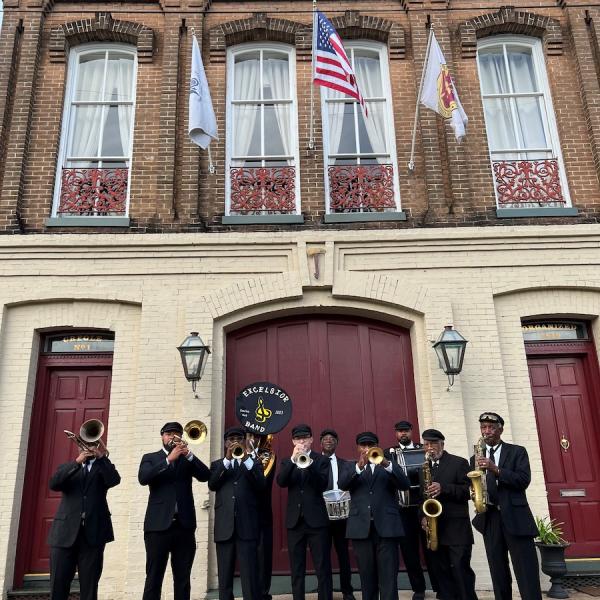 A band in uniforms plays in front of a two-story brick building.
