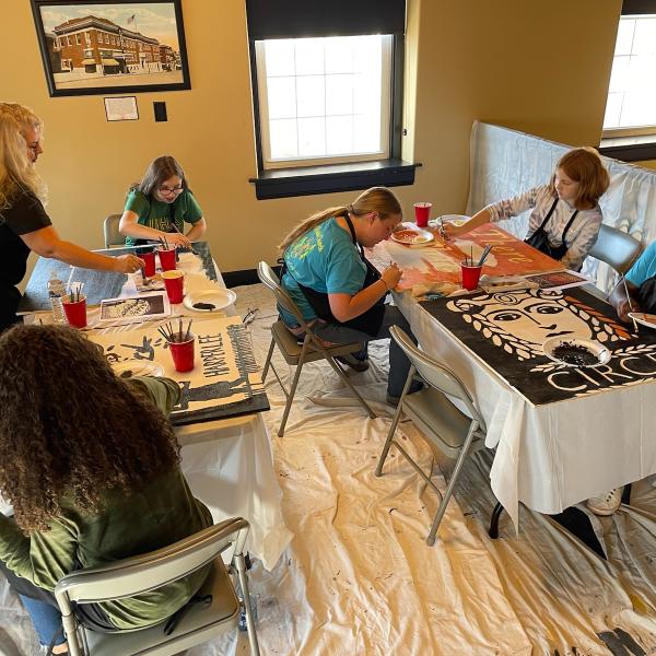 A group of 6 people sit around two tables painting book covers on large pieces of posterboard