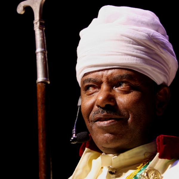 Black man wearing white turban and holding a cane with silver handle. 