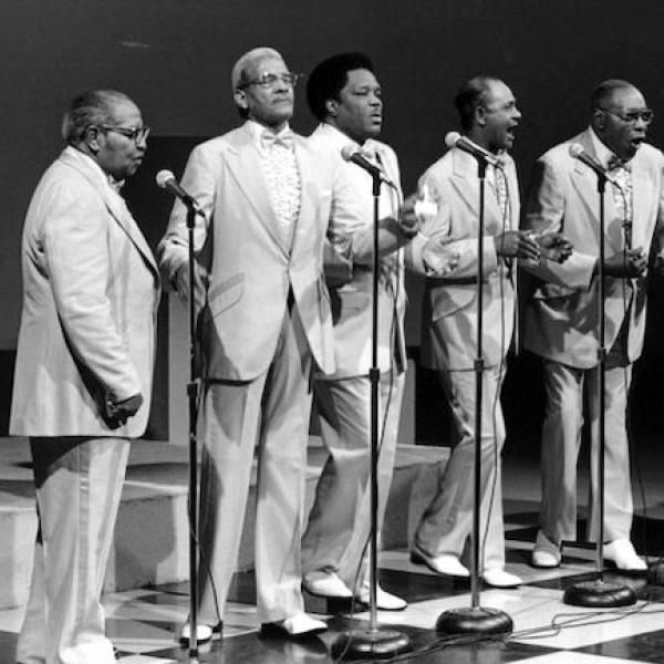 A group of men in suits sing from a satge.