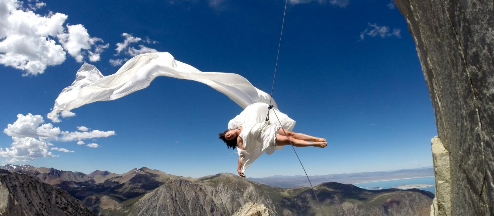Woman in flowing white dress dancing against side of cliff.