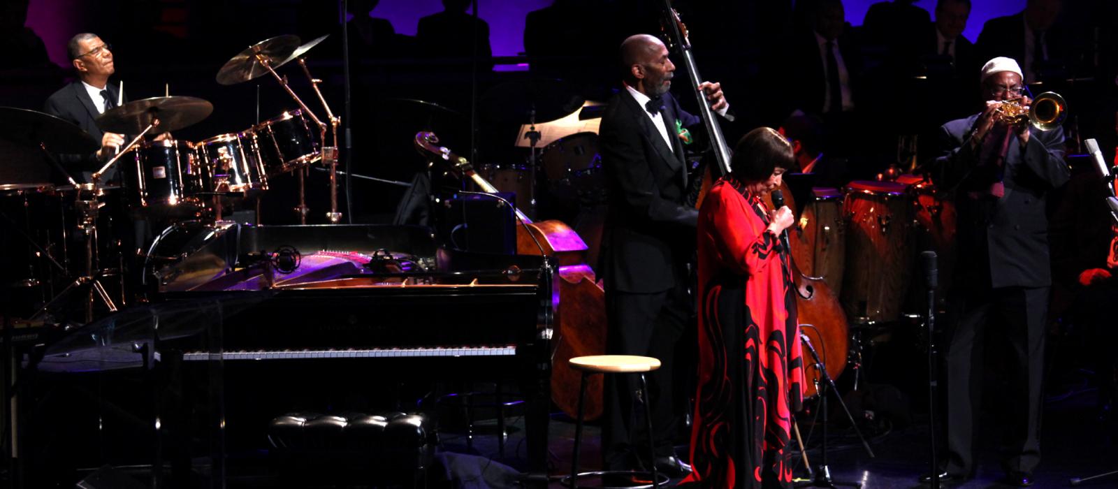 Group playing jazz onstage; from left, drummer, man playing bass, woman in red singing, and trumpet player.