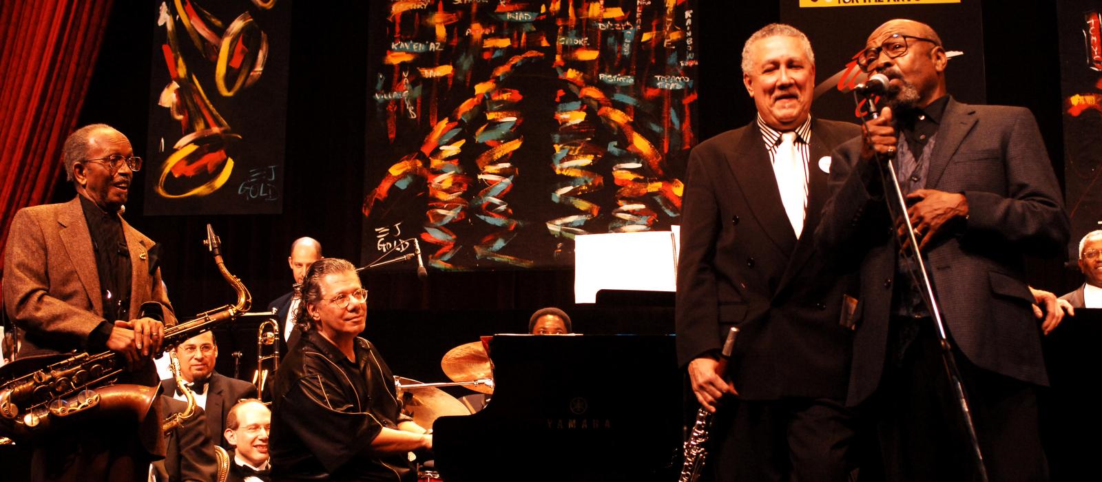 Group of men on stage playing jazz, saxophonist at far left, pianist, man holding clarinet, and man singing