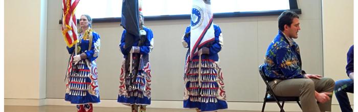 Three women in native american traditional dress holding the US and two native american flags on stage