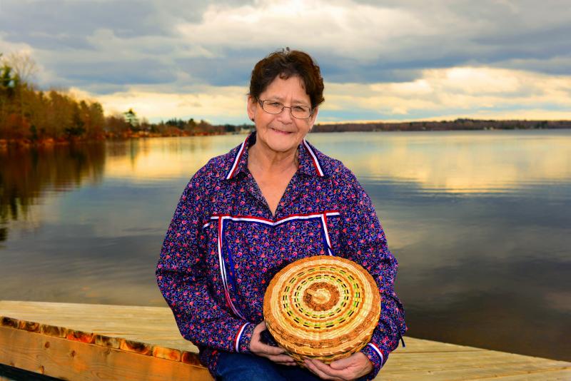 A woman stands in front of water, holding a woven basket