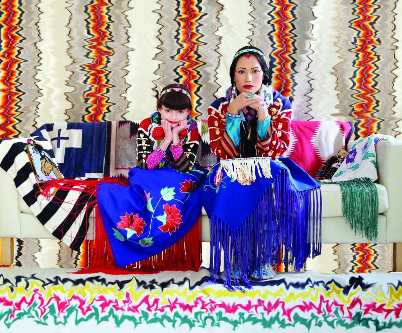 Woman and young girl dressed in Native garb sitting on a couch before a background of dizzying patterns.
