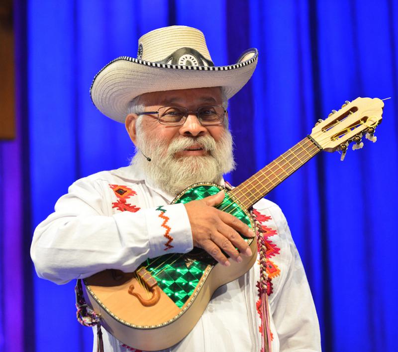 Musician with white beard in white suit and hat holding small guitar. 