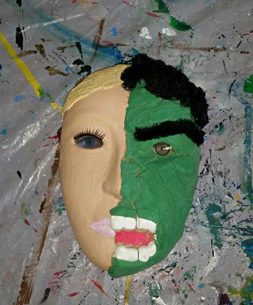 Mask against a swirling painted background. One side of face is flesh colored with painted blond hair, the other sie green with wool hair end eyebrow,, exaggerated teeth on one side