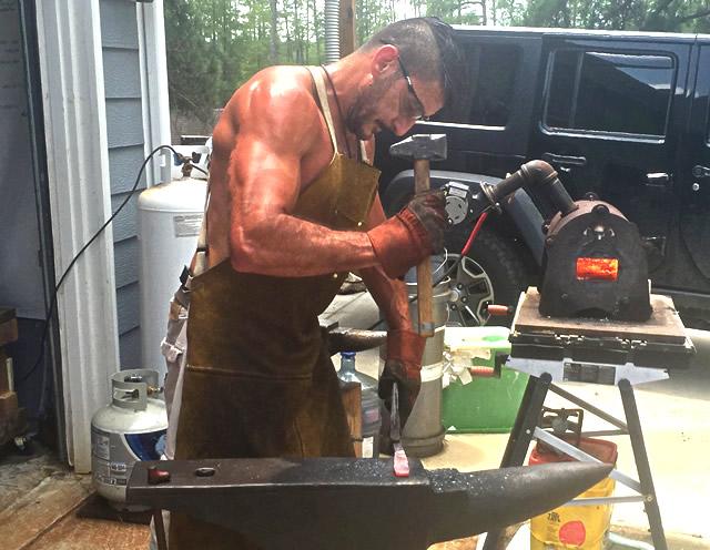 Man outdoors in his driveway hammering a hot piece of steel on an anvil