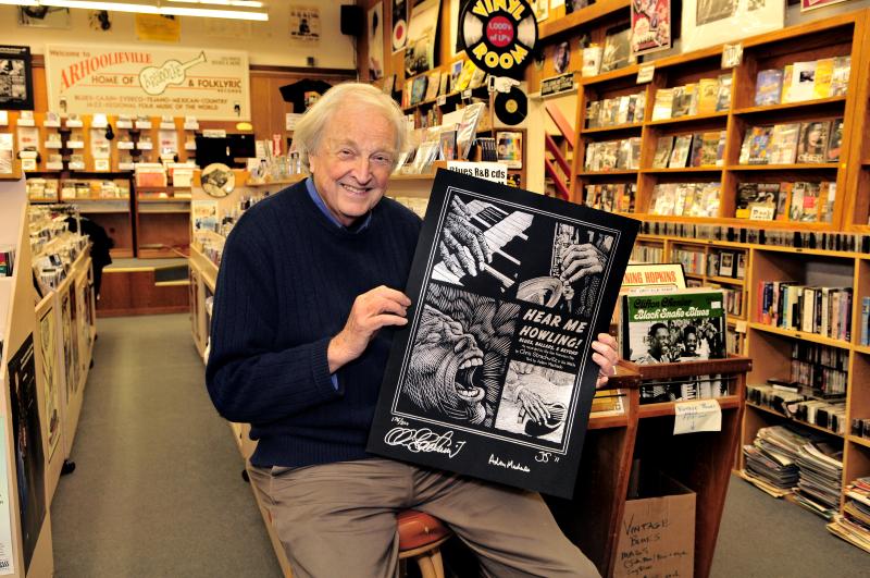 Man with white hair holding a poster sitting on a stool in a music shop with records and books. 