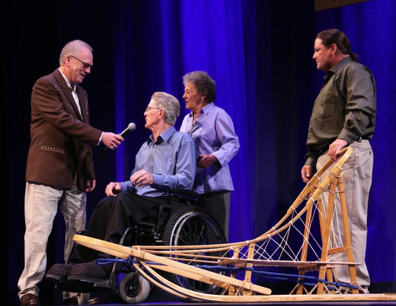 A man is holding a microphone toward a man sitting in a wheelchair, his wife standing behind him. In the foreground is a man standing next to a handmade dog sled.