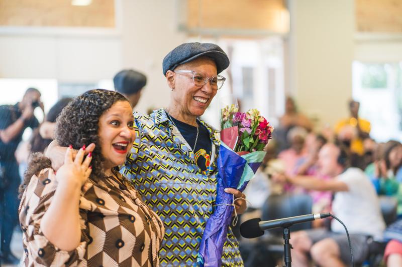 Black woman with long curly hair (left) smiling next to a Black woman with a hat on who is holding a bouquet of flowers (right).