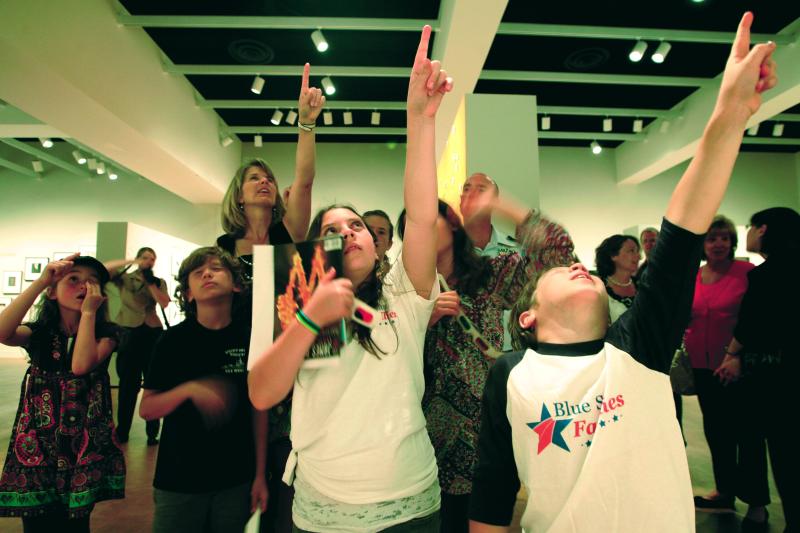 A group of children in a museum, all pointing and looking upward. 