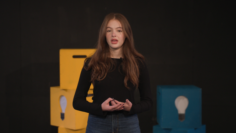 A young woman with long hair stands on a stage with several boxes painted yellow and blue behind her.