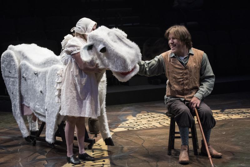 A man is seated on a stool, petting a large puppet of a cow. Another person dressed all in white hugs the cow.