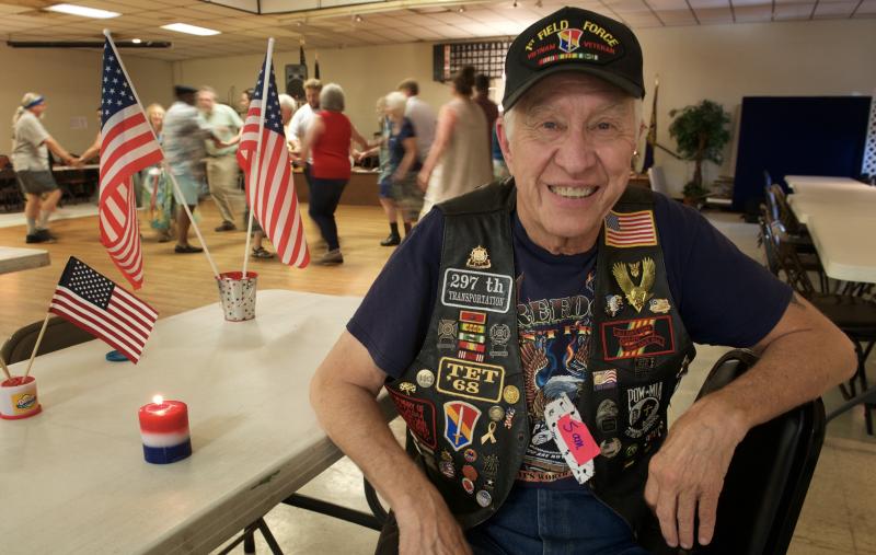 In a room with people square dancing, a smiling man wears a US Army 1st Field Force veteran’s ball cap and vest covered in military patches sits
