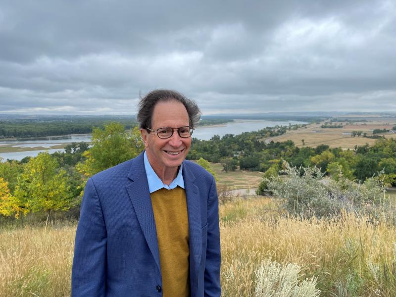Man wearing glasses on his face and a blue blazer, stands in the middle of natural scenery with various trees, a wheat field, and cloudy skies above. 