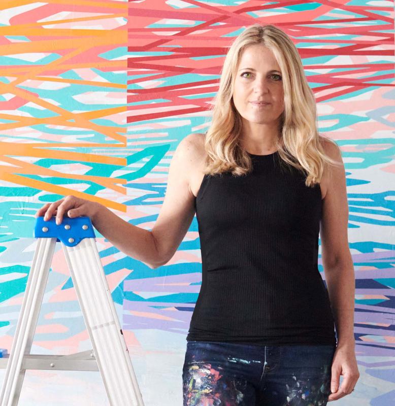 White woman in a black tank top and denim jeans holding on to a ladder, in front of a backdrop that has purple/orange/blue/green/red splatter paint. 