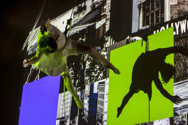 Woman held by ropes dancing on the side of a building with images projected on it. 