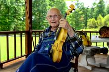 A man sits on a porch wearing overalls and holding a fiddle