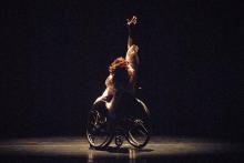 A dancer in a wheelchair lifts one hand upward as she performs onstage