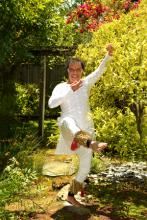 A man dressed in white balances on one leg in a garden.