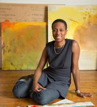 a young black woman with short hair sits on a wooden floor in front of two painted abstract canvases