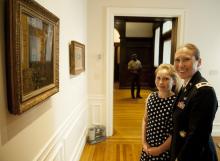 A white woman in military dress stands in front of two paintings with her young daughter