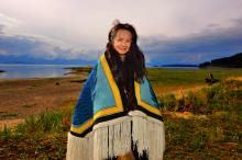 A woman stands on a shore wearing a woven blue and yellow blanket.