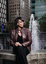 a black woman with glasses in black clothes and a brown leather jacket sitting on the edge of a fountain