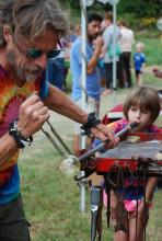 A young girl participates in a glass blowing demonstration with an artist helping.