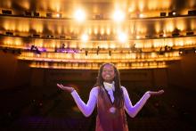 photo of young Black woman with long braids standing on stage with her back to audience and lights blazing behind her