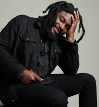 Man in black jacket and dreadlocks sits smiling with his head resting on one hand