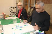 Two older men sit and paint at a table